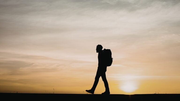 A solitary figure carrying a backpack while walking against a sunset sky.