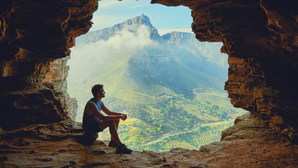 Un hombre se sienta en una cueva, contemplando un paisaje montañoso enmarcado por rocas, con una luz tenue y nubes a lo lejos.
